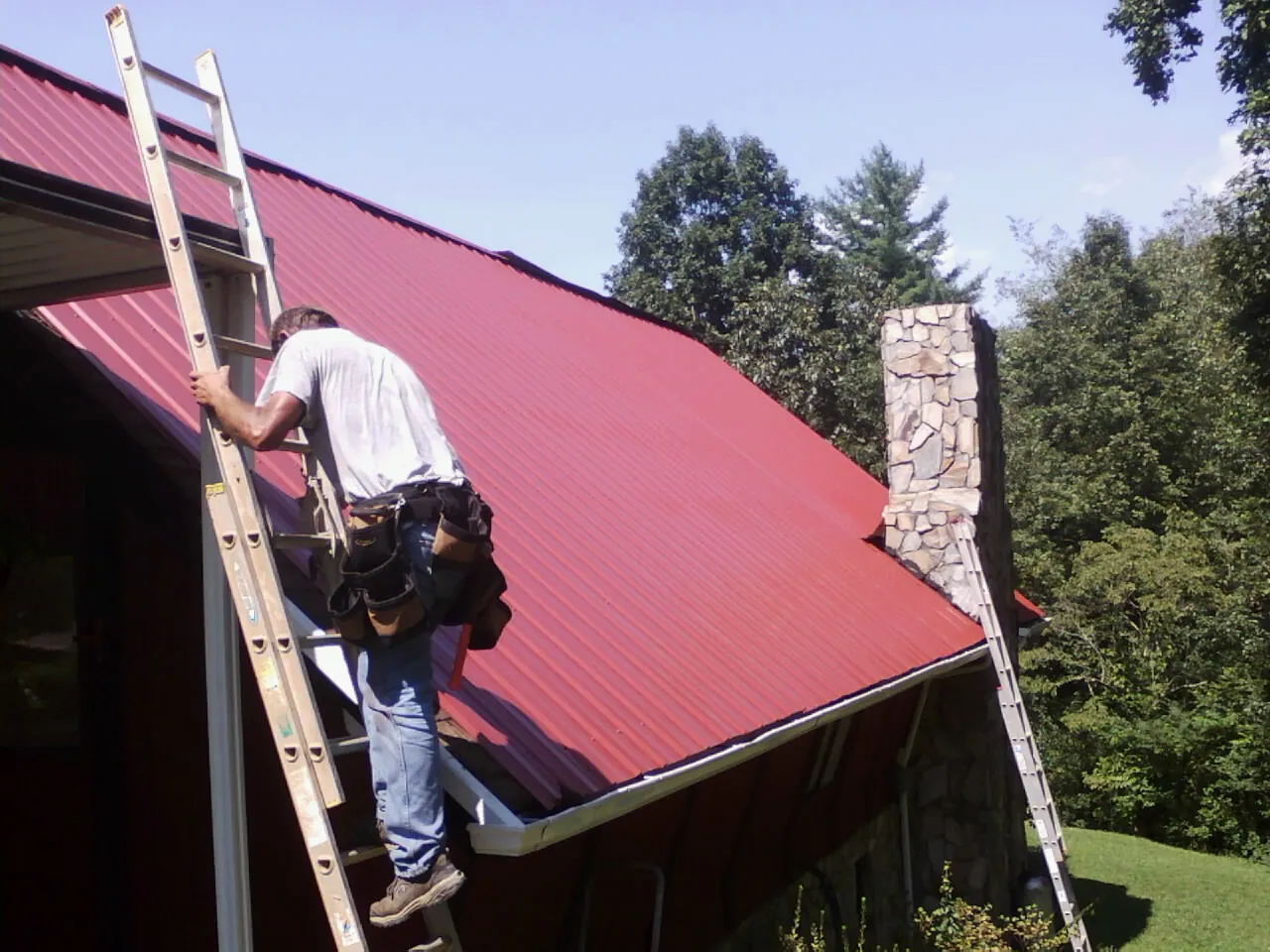 Roofer on a ladder beside a red metal roof and stone chimney — Soffit & Fascia Repair by Roofing Brothers in Metaline